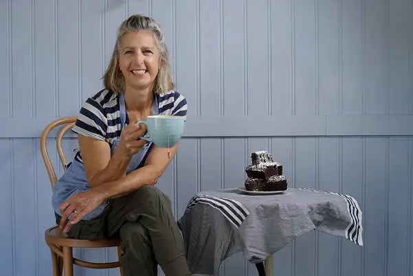 Woman smiling, holding a large cup, seated next to a table with chocolate cake.