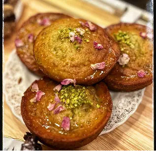 Four small cakes topped with pistachios and rose petals on a decorative plate.